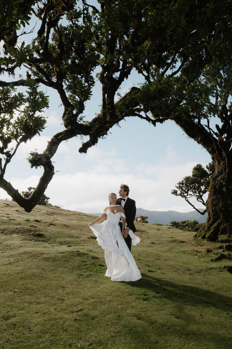 Brautpaar steht mit wehendem Kleid eng aneinander, editorial, steht vor Baum im Fanal Forrest, in Madeira