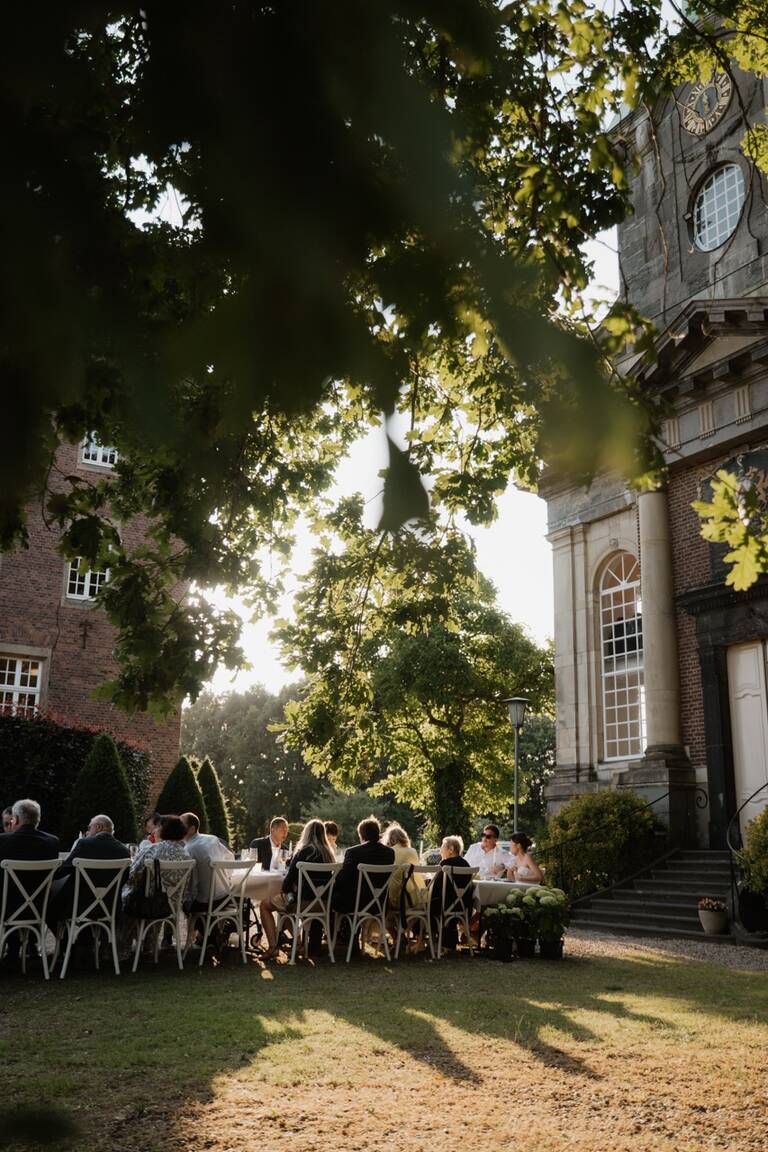 Sommerhochzeit, lange Tafel, bei der Hochzeitsgesellschaft zur goldenen Stunde draußen zu Abend isst, Schloss Diersfordt ist im Hintergrund zu sehen