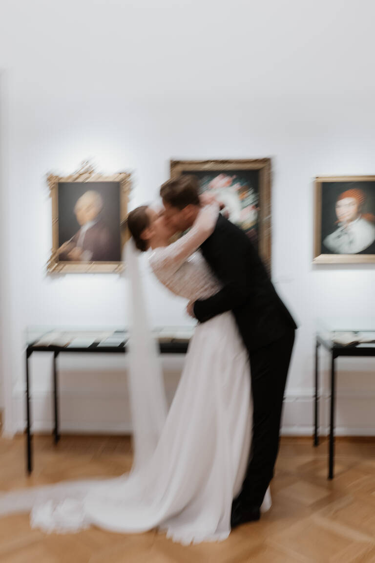Unscharfes Foto von intimer Szene, wie sich Brautpaar vor Kunst im Museum küsst